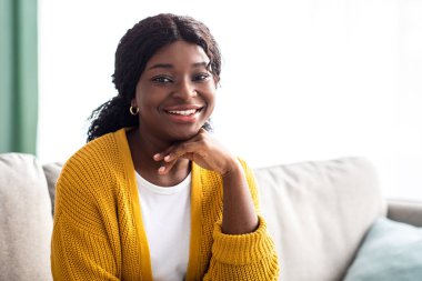 Evdeki kanepede poz veren çekici siyahi kadın, günlük kıyafetler giyiyor, eline yaslanıyor ve kameraya gülümsüyor, fotokopi çekiyor. Mutlu Afrikalı Amerikalı genç kadın hafta sonunu evde geçiriyor.