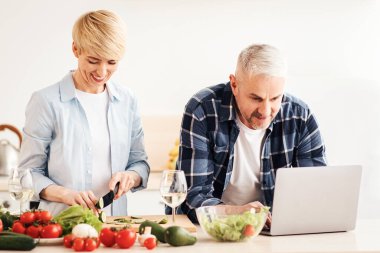 Mutlu orta yaşlı çift dizüstü bilgisayar kullanıyor, sağlıklı yemek, diyet, evde sebze salatası hazırlıyor, yemek tariflerini arıyor, internetten alışveriş yapıyor, mutfakta yemek kurslarını izliyor, panorama