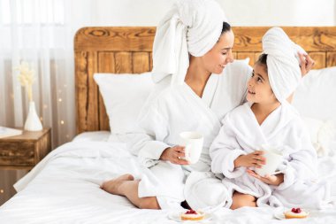 Oteldeki spa prosedürlerinden sonra küçük kızıyla çay içip kek yiyen genç bir anne. Kafası havluya sarılı, bornoz giymiş, evde yatağında oturan anne ve kadın çocuk.