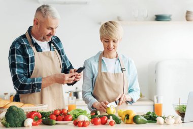 Birlikte lezzetli yemekler, sağlıklı yemekler ve vejetaryen yemekleri pişirin. Önlüklü mutlu bir son sınıf kadını yemek hazırlar, adam akıllı telefonuyla fotoğraf çeker mutfağın içinde parlak sebzeler ve meyve suyu ile.