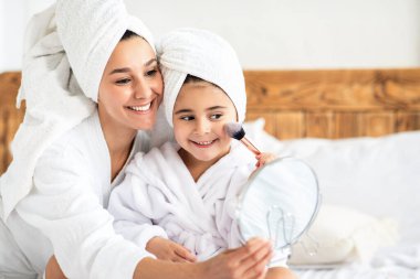 Neşeli küçük kız aynaya bakıyor, yanaklarını kızartıyor ve gülümsüyor. Bornozlu güzel anne ve kız evlerinde güzellik günü geçiriyorlar, yüzlerine makyaj yapıyorlar, uzay kopyaları yapıyorlar.