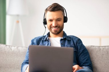 Hafta sonları film izleyen ya da Webinar 'a katılan rahat orta yaşlı bir adam, dizüstü bilgisayar ve kulaklık kullanıyor, oturma odasında kanepede oturuyor, evde boş zamanının tadını çıkarıyor, boşluğu kopyalıyor.