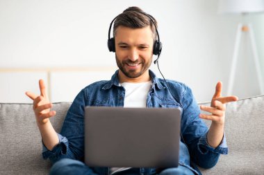 Olumlu sakallı adam evden video konferansı yapıyor, kulaklık ve laptop kullanıyor, konuşuyor ve el kol hareketi yapıyor. Keyifli orta yaşlı bir adam oturma odasındaki koltukta dinleniyor, görüntülü sohbet ediyor, boşluğu kopyalıyor.