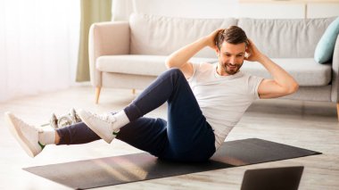 Spor kıyafetli orta yaşlı bir sporcu dizüstü bilgisayarın önünde mekik çekiyor, evden beden eğitimi dersi alıyor, içeriden oturma odası, fotokopi odası. Sportif sakallı adam günlük antrenman yapıyor.