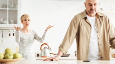 Tartışmalar, tartışmalar, skandallar, çift ilişkilerinde sorunlar ve kendini tecrit sırasında evde taciz. Kızgın orta yaşlı eş, beyaz mutfak iç dekorasyonunda üzgün yaşlı adama bağırıyor.