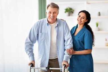 Walker 'ı kullanan mutlu yaşlı bir adamın portresi ve huzurevinde ona yardım eden genç bir hemşire. Olgun erkek. Bakıcı da yaşlı erkek. Huzurevine yürüyüşe gidiyor. Profesyonel yaşlı bakım servisi.