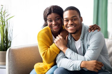Güzel siyahi aşıklar evde birlikte vakit geçirirken COVID-19 salgını, mutlu Afro-Amerikan çifti oturma odasında kanepede oturuyor, kameraya sarılıyor ve onlara bakıyor.