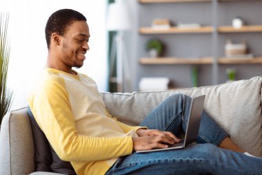 Uzaktan kumandalı işinden zevk alan mutlu siyah adam rahat oturma odasındaki kanepeye yaslanıyor, modern bir dizüstü bilgisayar kullanıyor ve gülümsüyor, yan görüş, kopyalama alanı kullanıyor. Serbest çalışma, uzak iş, ev konseptinden online çalışma