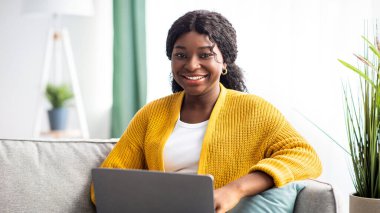 Evde çalışan örgülü süveter giyen neşeli genç siyahi kadın, kanepede oturuyor, dizüstü bilgisayar kullanıyor, fotokopi çekiyor. Rahat Afro-Amerikan bayan hafta sonunun tadını çıkarıyor, internette sörf yapıyor.