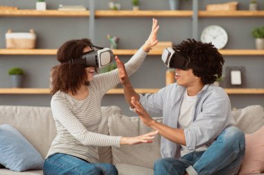 İnanılmaz bir eğlence, modern cihaz, evde birlikte eğlence. Mutlu binlerce yıllık Afro-Amerikan çifti VR gözlükleriyle kanepede oturuyor ve oturma odasının içindeki sanal dünyayı keşfediyor.