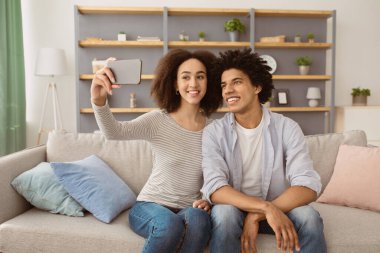 Moda, neşeli, pozitif insanlar selfie çekiyor. Gülümseyen Afro-Amerikan karısı ve kocası akıllı telefona bakıyor ve takipçilerle konuşuyor, sıcak oturma odasındaki kanepede oturuyor.