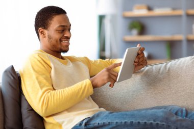 Oturma odasındaki kanepede dinlenen siyahi adam dijital tablet üzerindeki en yeni uygulamayı kullanıyor, uzayı kopyalıyor. Mutlu Afro-Amerikan erkeği evde eğleniyor, sosyal medyayı kontrol ediyor ya da arkadaşlarıyla sohbet ediyor.