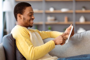 Evdeki koltukta dijital tabletle dinlenen neşeli siyahi adam, yan görüş, kopyalama alanı. Mutlu Afrika kökenli Amerikalı genç adam yeni mobil uygulamanın tadını çıkarıyor. Teknoloji ve ev eğlencesi