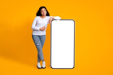 Gülümseyen Afrikalı Amerikalı kadın, büyük beyaz ekranlı akıllı telefona eğilip işaret ediyor uygulamanız ya da web siteniz için kopyalama alanı gösteriyor, sarı stüdyonun arka planında duruyor,