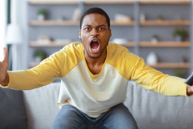 Öfkeli siyahi adam TV 'de futbol maçı izliyor, elinde uzaktan kumandayla bağırıyor ve el sallıyor, Afro-Amerikan genç adam futbol takımını kaybetmeye sinirlendi, iç mekan, fotokopi...