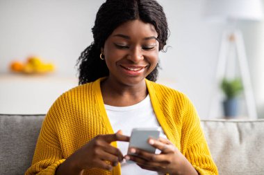 Modern akıllı telefon, ev içi ve fotokopi mekanı kullanan gündelik kıyafetli güzel siyahi bayana yakın çekim. İnternette blog okuyan ya da sosyal medyasını kontrol eden Afro-Amerikan genç bir kadın.