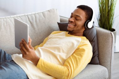 Neşeli siyah adam kanepede uzanıyor, evinde film izliyor, fotokopi çekiyor. Modern dijital tablet ve kulaklık kullanarak kanepeye yaslanan mutlu Afro-Amerikan genç adam.