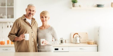 Pozitif duygular, neşeli ruh hali, günaydın, kahve molası, kahvaltı konforu ve ev konforu. Minimalist mutfak iç mekanında bir fincan sıcak içecekle gülümseyen yetişkin Avrupalı çift.