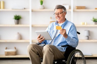 Tekerlekli sandalyedeki yaşlı adam kredi kartı ve tabletle yaşlılar yurdundan online alışveriş yapıyor. Neşeli kıdemli engelli erkek, elektronik para kullanarak internet mağazasından mal alıyor.