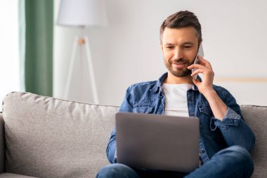 Bilgisayarlı, akıllı telefondan konuşan orta yaşlı sakallı adam oturma odasında koltukta oturuyor, fotokopi çekiyor. Mutlu adam serbest çalışıyor, müşterilerle telefon görüşmesi yapıyor.