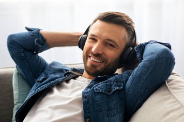 Kablosuz kulaklıklı, sakallı orta yaşlı bir adamın evde müzik dinlerken kameraya gülümseyen portresi. Mutlu adam yeni müzik seti kulaklığının keyfini çıkarıyor.
