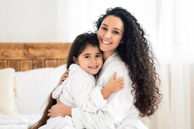 Bornozlu neşeli anne-kız yatak odasında sarılıyor, kadınlar gününü evde birlikte geçiriyorlar. Güzel, genç bir anne küçük kızını kucaklıyor. Hem gülümsüyor hem de kameraya bakıyor.