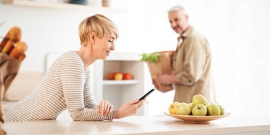 Covid-19 tecridi sırasında marketten taze organik sebze teslimatından geri bildirim. Uygulamada hizmet derecesi. Buzdolabının yanında kese kâğıtları olan mutlu bir koca, mutfakta telefonla konuşan bir kadın.