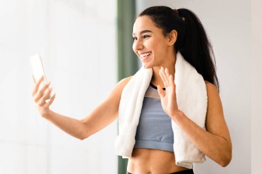 Spor giyimli genç bir bayan havluyla spor salonunda spor eğitmeniyle görüntülü görüşme yapıyor. Akıllı telefon ekranına el sallayan güzel bir bayan. Spor koçuna veda ediyor.