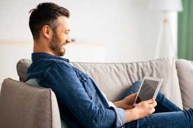 Gülümseyen sakallı adam boş ekranlı dijital tablet kullanıyor. Evdeki kanepede dinlenirken, yan görünüm, kopyalama alanı. Mutlu orta yaşlı bir adam yeni cep telefonu uygulaması kullanıyor.