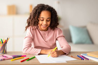 - Boş zaman. Mutlu Afro-Amerikalı kız renkli kalemler, renkli kalemler ve kalemlerle resim çiziyor, masada oturuyor ve gülümsüyor. Yaratıcı okul öğrencisi yeni hobisinin tadını çıkarıyor. Ev eğlencesi.