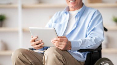 Tekerlekli sandalyedeki engelli yaşlı adamın evde online iletişim için tablet bilgisayar kullanması. Engelli erkek internette sörf yapıyor, içeride video izliyor, yakın plan çekiyor.