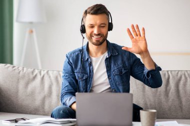 Gülümseyen sakallı iş adamı evden video konferansı yapıyor, kulaklık ve laptop kullanıyor, konuşuyor ve el kol hareketi yapıyor. Keyifli orta yaşlı bir adam oturma odasında koltukta oturuyor, görüntülü konuşma yapıyor, boşluğu kopyalıyor.