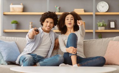 Covid-19 salgını ve insani duygular sırasında evde eğlence. Heyecanlı genç Afro-Amerikalı kadın televizyonda parmağını gösteriyor, adam uzaktan kumandayı değiştiriyor, oturma odasındaki kanepede oturuyor.