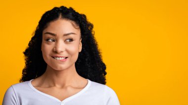 Güzel teklif. Meraklı siyah bir kadın ilgisini başka bir yere çekiyor, pozitif Afrikalı Amerikalı bir kadın dudaklarını ısırıyor ve gülüyor, sarı arka planda tek başına duruyor, Panorama
