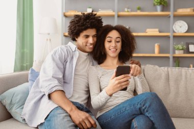 Covid-19 sırasında evde alışveriş yapmak, komik video, online toplantı ve internette sörf. Mutlu Milenyum Afrikalı Amerikalı erkek ve kadın telefona bakar, oturma odasındaki kanepede oturur.
