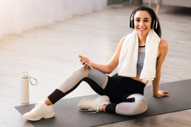 Kablosuz kulaklıklı ve akıllı telefonlu formda bir bayan mola veriyor, spor yaparken spor salonunda oturuyor, su içiyor, kameraya bakıyor ve gülümsüyor, boşluğu kopyalıyor.