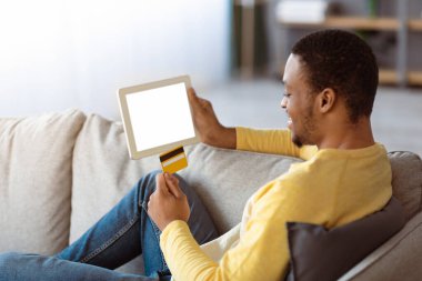 Kredi kartı ve dijital tabletli Afrikalı Amerikalı genç adam evde boş ekranlı koltukta oturuyor, alışveriş yapıyor veya online bankacılık yapıyor, mobil uygulama kullanıyor, kopyalama alanı kullanıyor, modelleme yapıyor.
