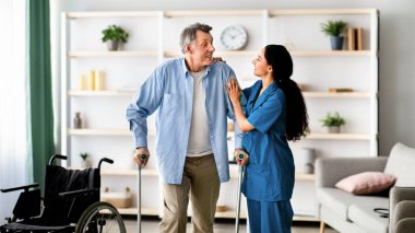 Olumlu bayan hemşire yaşlı erkeğin koltuk değnekleriyle yürümesine yardım ediyor. Kıdemli adam içeride bakıcıdan yardım alıyor. Engelli yaşlı hastalar için profesyonel tıbbi bakım