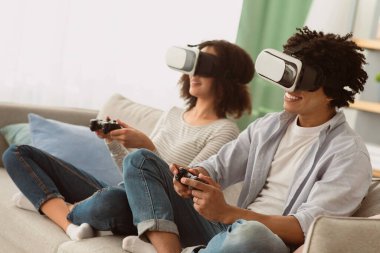 Evde teknoloji, birlikte eğlence ve covid-19 'da kendini izole etme. Gülümseyen bin yıllık Afro-Amerikan kadın ve erkek oturma odasındaki koltukta oturuyorlar. VR gözlükleriyle, joysticklerle oyun oynuyorlar.