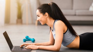 Sportif genç bayanın spor minderinde dizüstü bilgisayar kullanarak fotokopi çekerken yan görüntüsü. Spor giyim sektöründe mutlu bir kadın internette koltuk değnekleriyle antrenman yapmayı seçiyor. Spor eğitimi, sınıf çevrimiçi