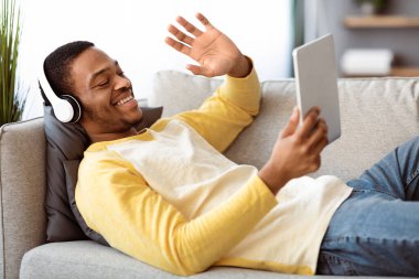 Neşeli Afrikalı Amerikalı genç adam arkadaşlarıyla veya sevgilileriyle video konferansı yapıyor, dijital tablet ve kablosuz kulaklık kullanıyor, tablet ekrana bakıyor, gülümsüyor ve el sallıyor, ev içini kopyalıyor.