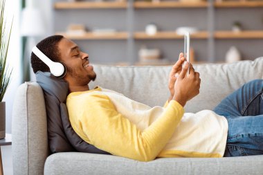 Rahat siyah adam kanepede uzanıyor, evinde film izliyor, fotokopi çekiyor. Mutlu Afrikalı Amerikalı genç adam kanepeye yaslanıyor, modern dijital tablet ve kablosuz kulaklık kullanarak, yan görüş