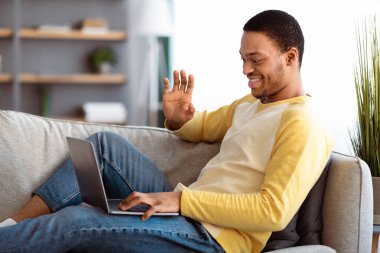 Neşeli siyah adam dizüstü bilgisayara el sallıyor, kanepeye yaslanıyor ve arkadaşlarıyla veya sevgilileriyle görüntülü görüşme yapıyor, ev içi, yan görüş, kopyalama alanı. COVID-19 salgını sırasında iletişim