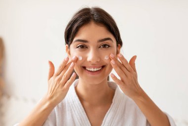 Yüzüne krem süren neşeli kadın banyodaki kameraya gülümseyen cilt nemlendirici. Yüz Cilt Kozmetiği, Kadın Güzellik Programı