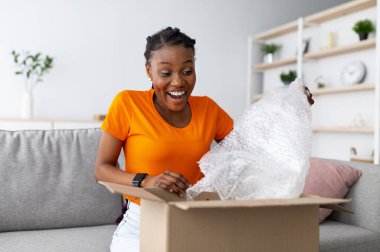 Karton paketleri açan mutlu siyahi kadın, başarılı alışverişe duygusal tepki veriyor, plastik ambalajı çıkarıyor, evden büyük alışverişlerle tatmin oluyor. Çevrimiçi mağaza ve dağıtım kavramı