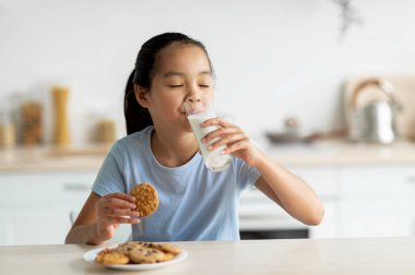 Asyalı kız süt içiyor ve kurabiyelerden hoşlanıyor, mutfakta oturuyor ve abur cubur yiyor, boş yer. Tatlı çocuk taze pişmiş bisküvi ve kalsiyum içeceği ile bir ısırık alıyor.