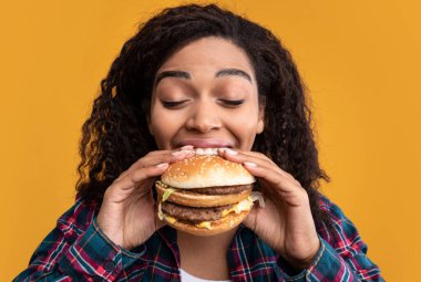 Oburluk. Komik, Afro-Amerikan, Hamburger Yiyen Kadın portresi. Kıvırcık saçlı, kıvırcık saçlı ve lezzetli fast food yiyen, turuncu stüdyo arka planında izole edilmiş siyah kadın.