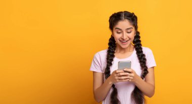 Halk ve Teknoloji Konsepti. Gülümseyen genç Hintli kadının portresi cep telefonunu kullanıyor, arkadaşlarıyla internette sohbet ediyor, sarı stüdyo arka planında, afişte, bedava fotokopi alanında tek başına duruyor.