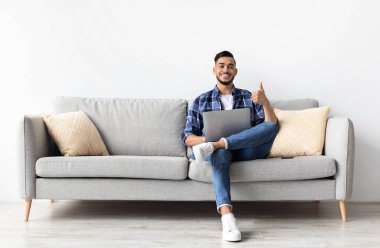 Hoşuma gitti. Mutlu Orta Doğulu Adam 'ın portresi kanepeye oturmuş dizüstü bilgisayarla işaret dilini gösteriyor, el hareketlerini gösteriyor, çalışıyor ya da uzaktan çalışıyor oturma odasındaki kameraya poz veriyor, tüm vücut uzunluğu...