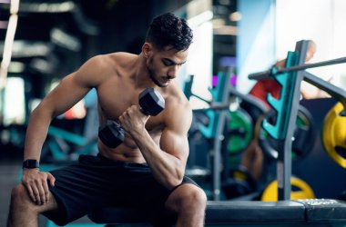 Modern spor salonunda dambıllarla egzersiz yapan, kaslı, güçlü ve kaslı Arap genç adam, kas gücü için ağır ağırlıklarla antrenman yapıyor, serbest alan yaratıyor.